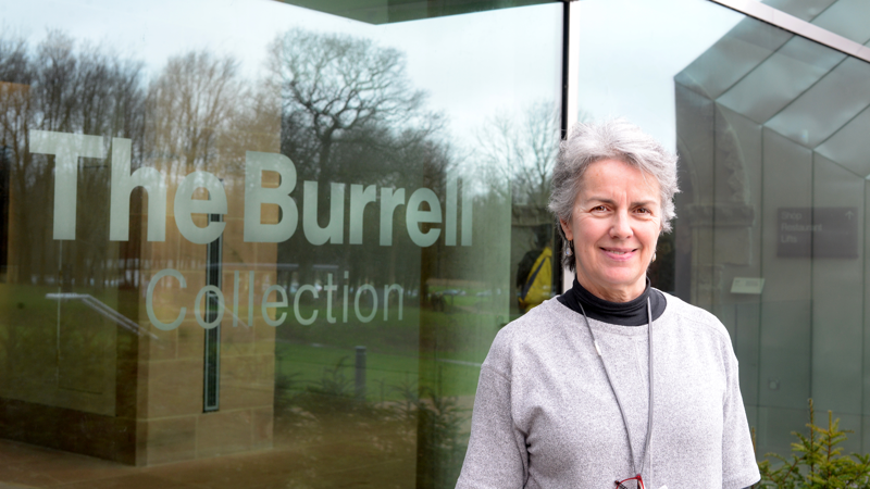 A person standing outside the Burrell collection. They have grey hair and a grey jumper with a lanyard around their neck. They are smiling at the camera.
