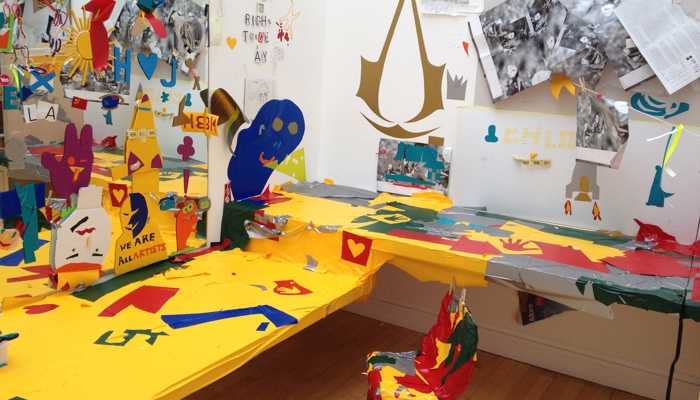 A vibrant, collage-filled art room featuring a brightly decorated table and chair surrounded by colourful cut‑outs, bold text, and expressive mixed‑media artwork on the walls.