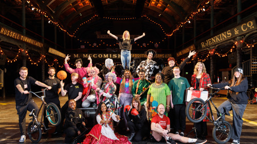 A group of people stand in a large indoor space dressed in brightly coloured costumes and sportswear with some holding sport equipment including a basketball and BMX bikes