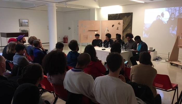 A gallery talk in a white-walled space, with an audience seated in rows facing a panel of speakers at a table, with artworks and a projected image behind them.