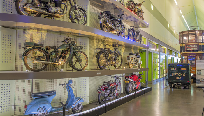 Image of a wall of vintage motorised bikes. The wall is in Riverside museum, the room has a green tint to the walls and it is well lit. In the background you can see other vintage vehicles, like a bus and car.