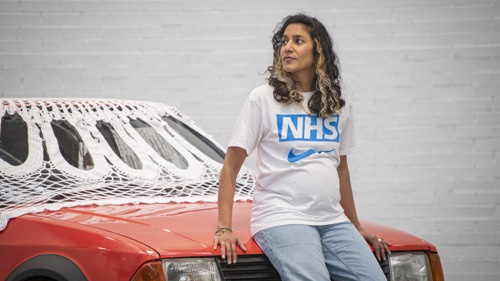 artist with brown hair and blonde highlights wearing NHS t-shirt sitting on the hood of an orange car