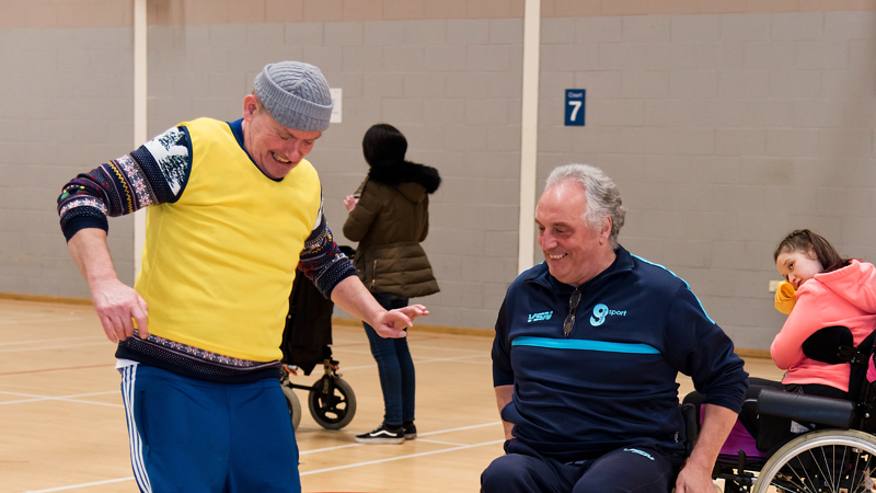 Two people smiling while playing indoor football with a large ball, one of the people is a wheelchair user 