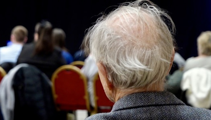 A close up of an older person from behind sitting and watching a performer singing on stage. 