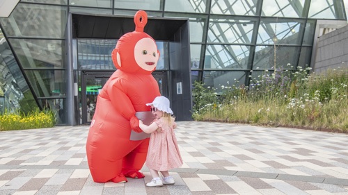 A person in a large red inflatable character costume is holding hands with a young child wearing a sun hat in front of a large glass convention centre
