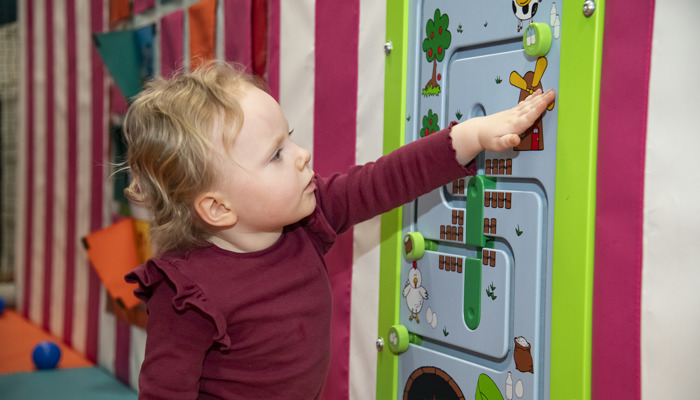 A young child pointing and looking at a colourful puzzle of farm animal on the wall of a soft play area.