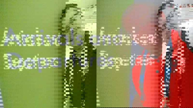 Andrew McVie wearing his red Clyde-sider uniform in front of the arrivals and departures sign at Glasgow Airport during the 2014 Commonwealth Games.