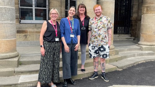 A close up of a group of four people standing on the steps outside a stone library building. They are all smiling and wearing the same pink lanyards.