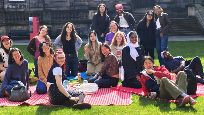 Group of young people sitting and standing on picnic blankets on a grassy lawn in front of a large stone building (Kelvingrove Art Gallery), posing together outdoors on a sunny day with snacks, drinks, and bags visible around them.