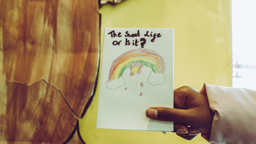 A hand holds a drawing with a rainbow with the questions "it's a sweet life, or is it"