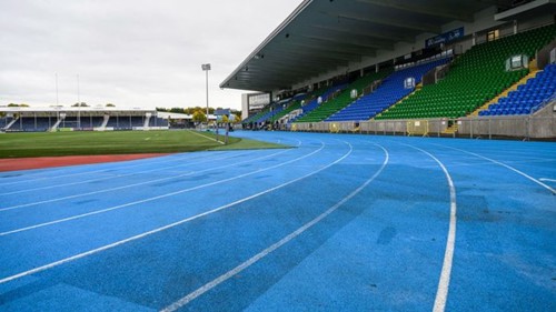 Section of a blue outdoor running track in a stadium. There are large tiered seating areas to the sides and a grassy area in the middle of the track.