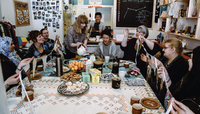 Many community members sit around a large table laughing and sharing stories. There are cakes, tea and treats that are being served to everyone. 