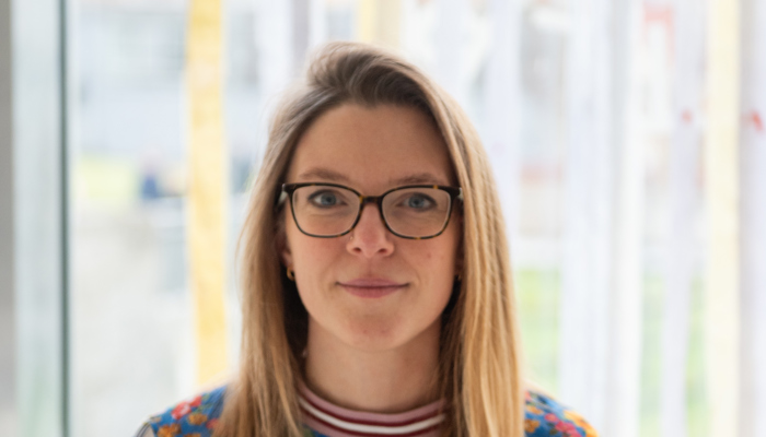 A person with glasses and long blonde hair stands in front of a window smiling. 