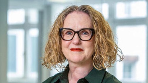 A headshot of Susan Deighan, a person with curly red hair, wearing glasses, a green shirt and beaded wooden necklace