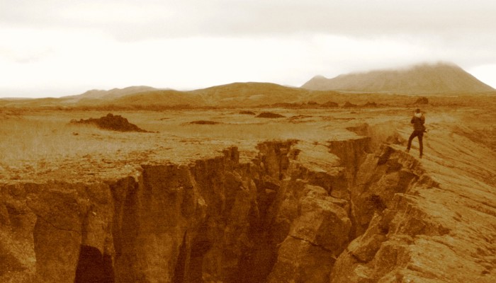 Sepia photograph of a person standing at the edge of a deep rocky fissure in a rugged landscape