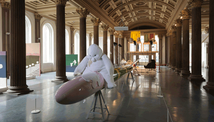 A large, playful sculpture of a soft, white, cartoon-like figure sits atop a long, glossy rocket-shaped form in a grand columned gallery, surrounded by colourful artworks and display structures.