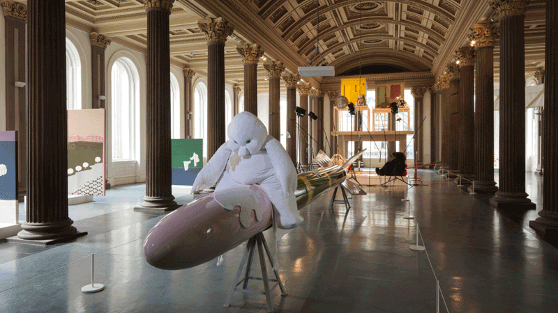 A large, playful sculpture of a soft, white, cartoon-like figure sits atop a long, glossy rocket-shaped form in a grand columned gallery, surrounded by colourful artworks and display structures.