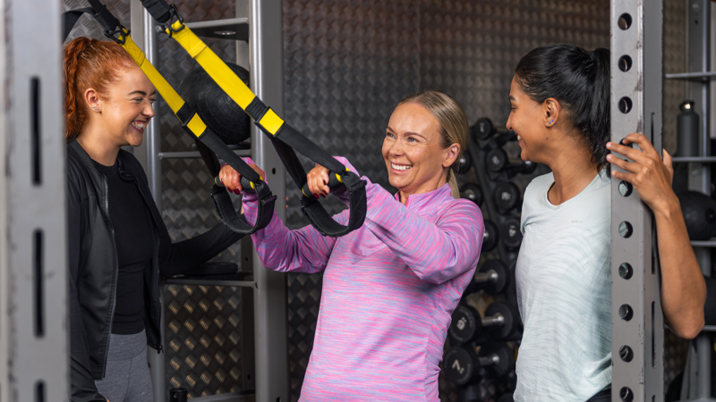 Three adults smiling while standing at a weights rack in the gym, the person in the middle is holding onto yellow and black TRX straps.