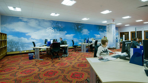 A room in the Family History centre at The Mitchell Library featuring multiple desks with PCs and bookcases, there are a few people sitting at the desks looking at the computer screens.