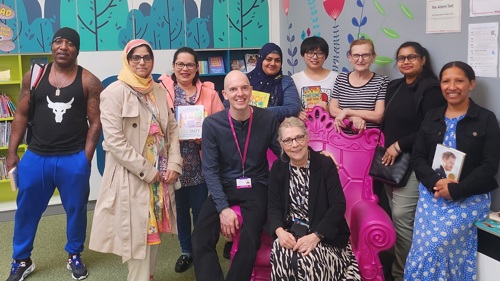 A group of ten people are gathered in a library for a group photo, two of them are sitting on a large plastic purple throne, while those standing behind each hold a book in their hands