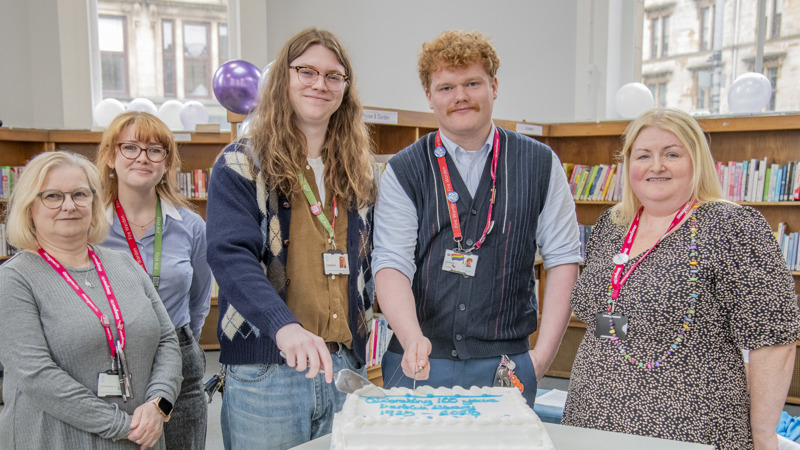 A group of five people gathered around a large white cake to celebrate the 100 anniversary of the library they are standing in