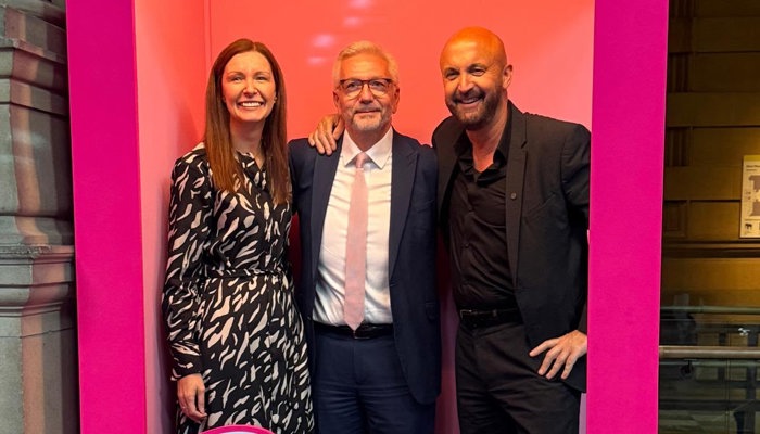 Three people are standing and smiling inside a large pink doll box to promote the Barbie exhibition