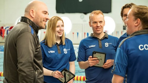 A group of five colleagues in the same blue t-shirts having a conversation at the side of an indoor pool. Two of them have tablet computers in their hands