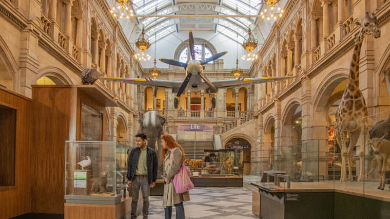 The interior of Kelvingrove museum in Glasgow, which shows a display hall with a high glass ceiling, ornate stone arches, and hanging chandeliers. A historic aircraft is suspended from the ceiling above the central space. In the foreground, two visitors stand beside a glass display case containing taxidermy animals, while additional animal exhibits, including a giraffe, are visible along the sides. The hall is brightly lit, with patterned stone flooring and display cases arranged throughout.