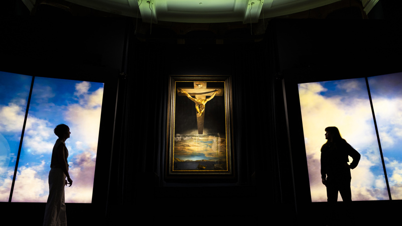 Visitors view Salvador Dalí's Christ of Saint John of the Cross at Kelvingrove Art Gallery and Museum, displayed between illuminated panels showing clouds and sky.