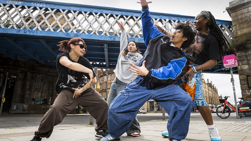 Four street dancers perform expressive, energetic moves beneath a blue iron bridge in an urban area. They wear casual, loose-fitting clothes and sneakers, striking dynamic poses that convey rhythm and creativity. Behind them, stone buildings and a pink sign reading “People Make Glasgow” highlight the lively city backdrop.