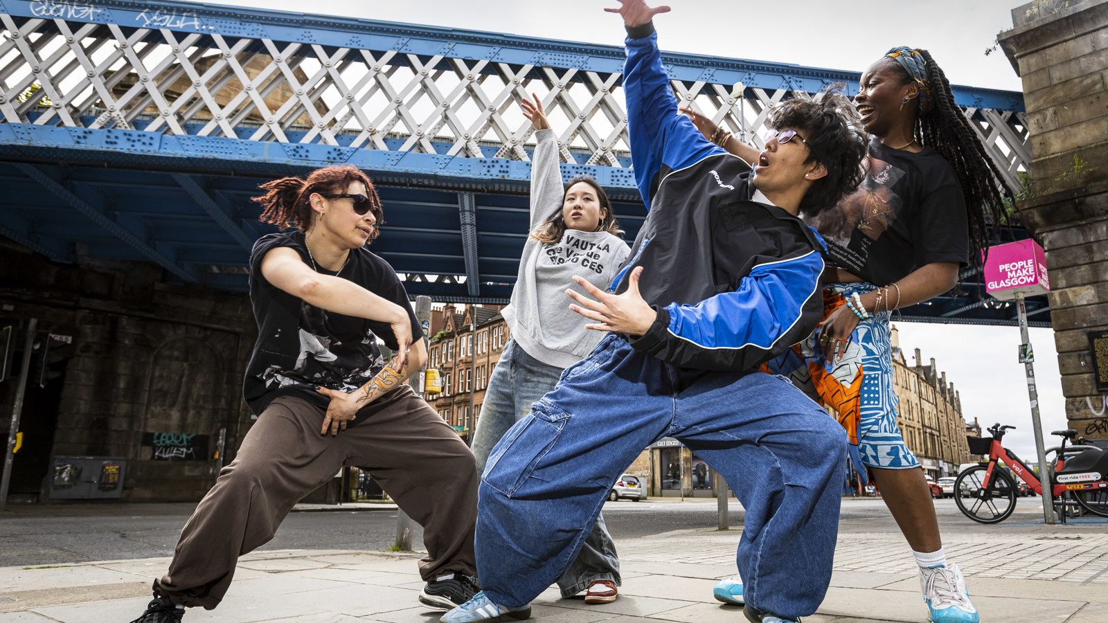 Four street dancers perform expressive, energetic moves beneath a blue iron bridge in an urban area. They wear casual, loose-fitting clothes and sneakers, striking dynamic poses that convey rhythm and creativity. Behind them, stone buildings and a pink sign reading “People Make Glasgow” highlight the lively city backdrop.
