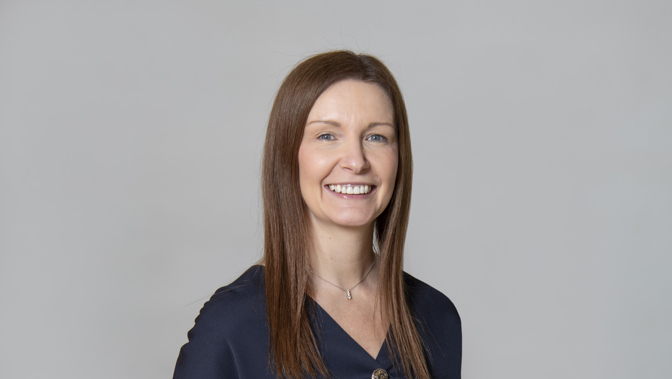 Studio headshot of a person with long brown hair, facing forward against a plain light grey background, wearing a dark navy top and a small necklace, with a neutral posture and visible teeth.