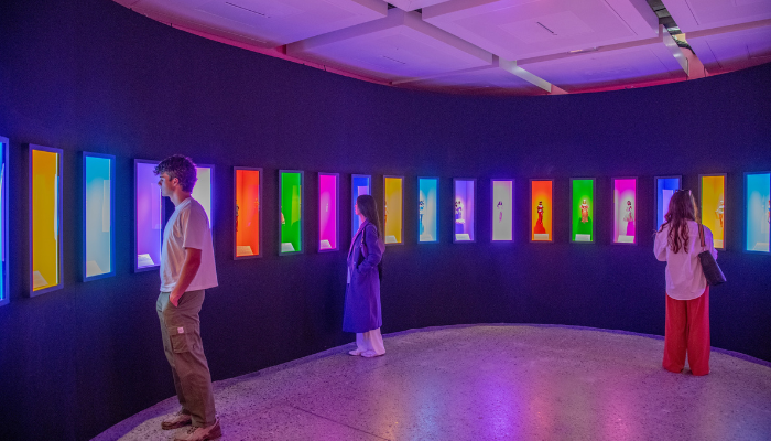 3 visitors in wide space looking at a curved wall with neon blocks embedded in the wall containing Barbie dolls