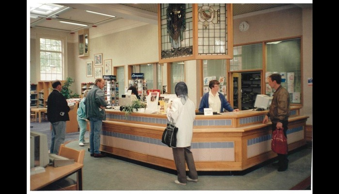 Photograph of the counter of a library. There are staff behind the desk and customers in front.