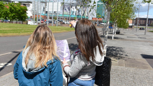 Two people are facing away holding an old photo of Celtic Park whilst looking at the building.