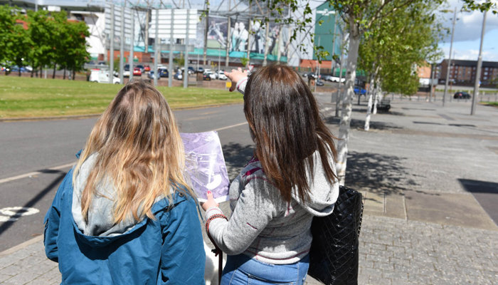 Two people are facing away holding an old photo of Celtic Park whilst looking at the building.