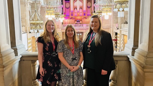 Three people standing in the upper gallery of a large sandstone and marble museum. They are all wearing the same pink lanyard
