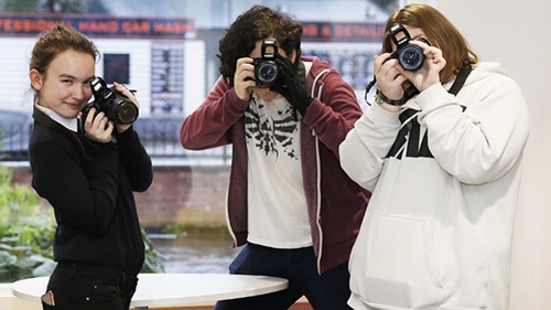 Three people hold cameras to their faces and pretend to take a picture of the person who is photographing them. 