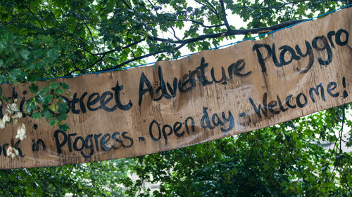 A sign hanging between trees and leaves which says Baltic Street Adventure Playground.