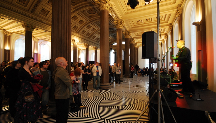 A crowd gathers in an ornate, columned hall with a patterned black-and-white floor, watching a live music performance on a small stage lit by warm lights