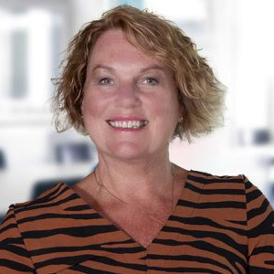 A headshot of Jan Buchanan, a person with blonde wavy hair wearing a brown and black striped top
