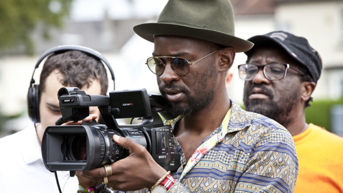 A person with sunglasses and a hat films on a professional camera.