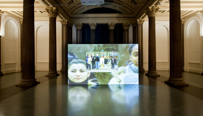 A large, ornate gallery hall with a high, arched ceiling richly decorated with detailed moulding and patterned panels. A wide rectangular screen sits directly on the floor, displaying a video image with close ups of young people’s faces a small screen suspended in front shows a small group of people standing together outdoors. The space is evenly lit with warm overhead lighting, highlighting the classical features of the room while the contemporary video installation becomes the focal point in the gallery.