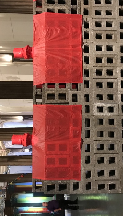 A large installation of stacked concrete blocks in a grand columned hall, with two red fabric-covered forms draped over the structure and colourful washing line of clothes and other objects hanging below the ornate ceiling behind. 