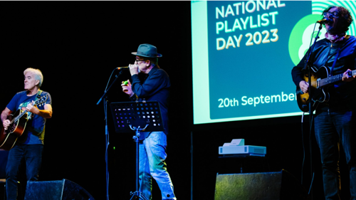 Three musicians performing on a stage, two are playing guitars, the other a mouth organ