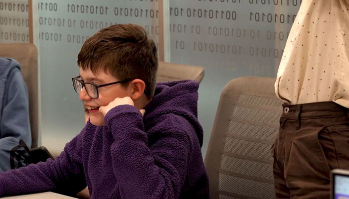 Young person with glasses using a computer in a learning session, seated at a desk in a library.