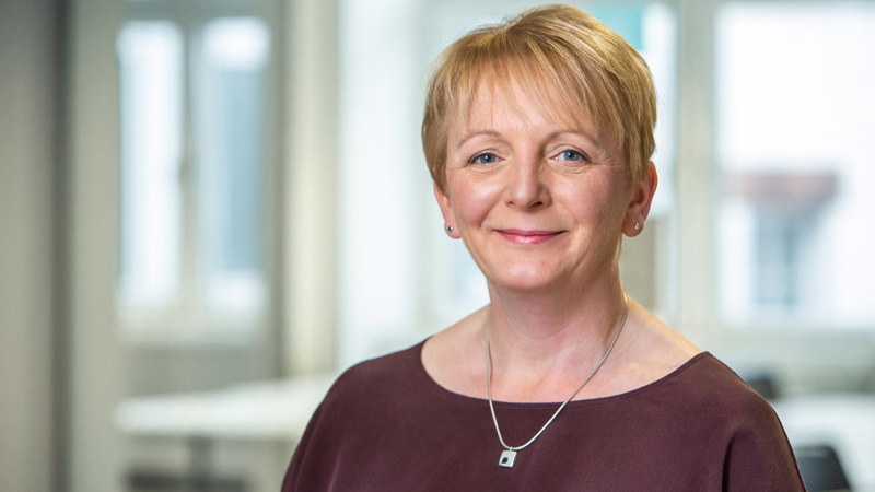 Headshot of a person with short blonde hair, wearing  brown top and a silver necklace, smiling in front of a window in an open plan office space