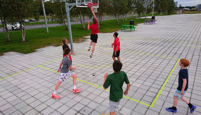 A young person in red jumps to shoot a basketball while six others stand around the hoop.
