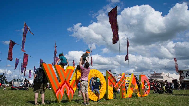 Children and adults playing on a large, colourful WOMAD sign in a grassy field under a bright blue sky.