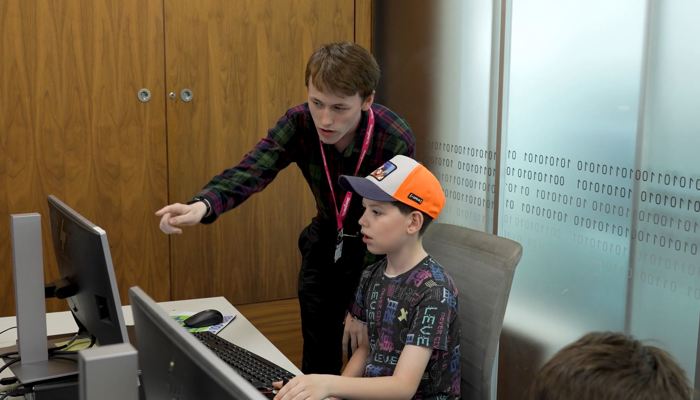 A volunteer points to a computer screen while supporting a young person during a coding activity.